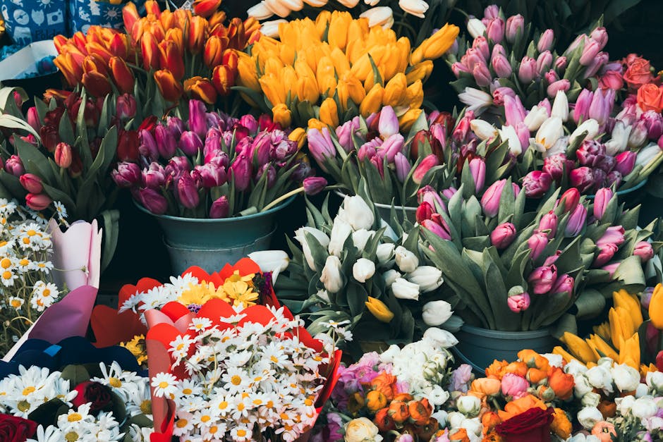 A vibrant display of fresh flowers including yellow tulips, white and pink tulips, and red, orange, and white roses arranged in blue buckets and bouquets. The flowers are densely packed, showcasing a