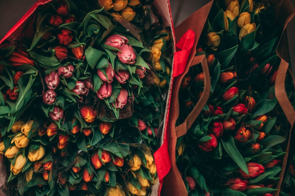 Multiple bouquets of fresh tulips in shades of pink, yellow, red, and orange with green leaves, wrapped in transparent plastic and brown paper, arranged inside open florist delivery bags. The tulips a