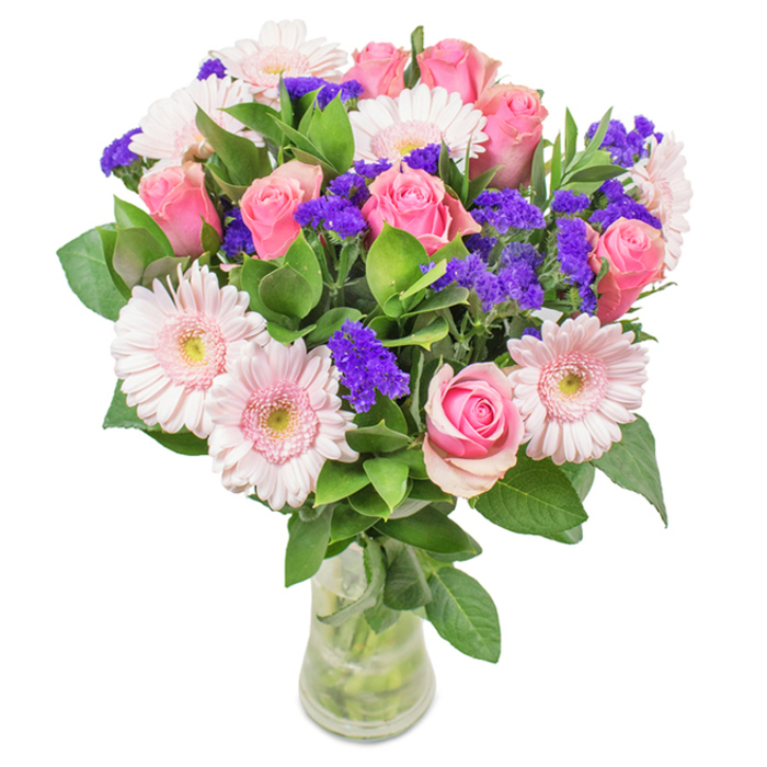 A striking, thoughtfully assembled flower arrangement sits in a clear glass vase, the blooms radiant against a pure white background-a nod to the vibrancy of Hackney Marshes. The heart of the bouquet features between seven and nine classic pink roses, their petals ranging from tightly wound buds to gently opening spirals, showcasing textures that evoke both tradition and freshness. Flanking them are five to seven pale pink gerbera daisies, each with broad heads and slightly lighter centres, their symmetry and open faces reflecting both cheerfulness and poise. Sprays of bright purple statice weave throughout, their colour reminiscent of wildflowers by the marsh, bringing lively contrast without overwhelming the bouquet's gentle palette. An abundance of deep green foliage wraps the base, fresh and full, mirroring the lushness of Hackney's green corridors. The transparent vase, revealing tangle-free, healthy stems and fresh water, completes the look for authentic flower delivery. Perfect for birthdays, anniversaries, or simply brightening a Mare Street kitchen table, this expertly balanced bouquet expresses the creative heart and welcoming warmth of Hackney Marshes.